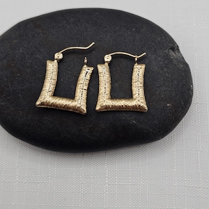 May include: A pair of gold-colored, square hoop earrings. The earrings have a textured surface and a classic design. The earrings are displayed on a black stone against a white background. Jewelry.