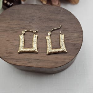 May include: A pair of gold-colored, square hoop earrings. The earrings have a textured surface and a post-and-loop closure. The earrings are displayed on a wooden surface.
