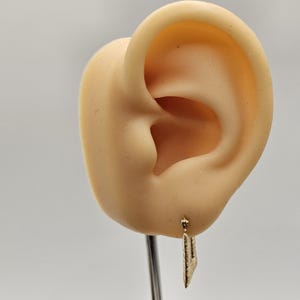 May include: A gold-colored earring with a textured, leaf-like design hangs from a mannequin ear. The earring is suspended from a small hook. The ear is a light beige color, and the background is a neutral gray.