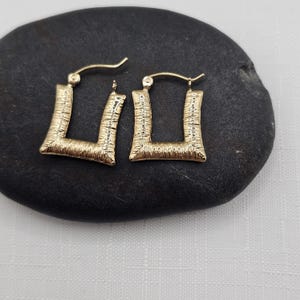 May include: A pair of gold-colored square hoop earrings. The earrings have a textured surface and a post closure. The earrings are displayed on a black stone surface.