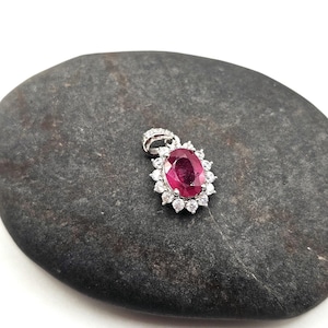 May include: A silver pendant with a large oval ruby gemstone at the center, surrounded by smaller, round, clear gemstones. The pendant is set on a dark gray, smooth stone surface.