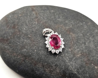 May include: A silver pendant with a large oval ruby gemstone at the center, surrounded by smaller, round, clear gemstones. The pendant is set on a dark gray, smooth stone surface.