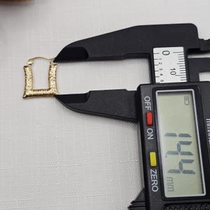 May include: A gold-colored, square-shaped earring is measured with a black digital caliper. The earring has a textured surface. The caliper displays a measurement of 14.4 mm.
