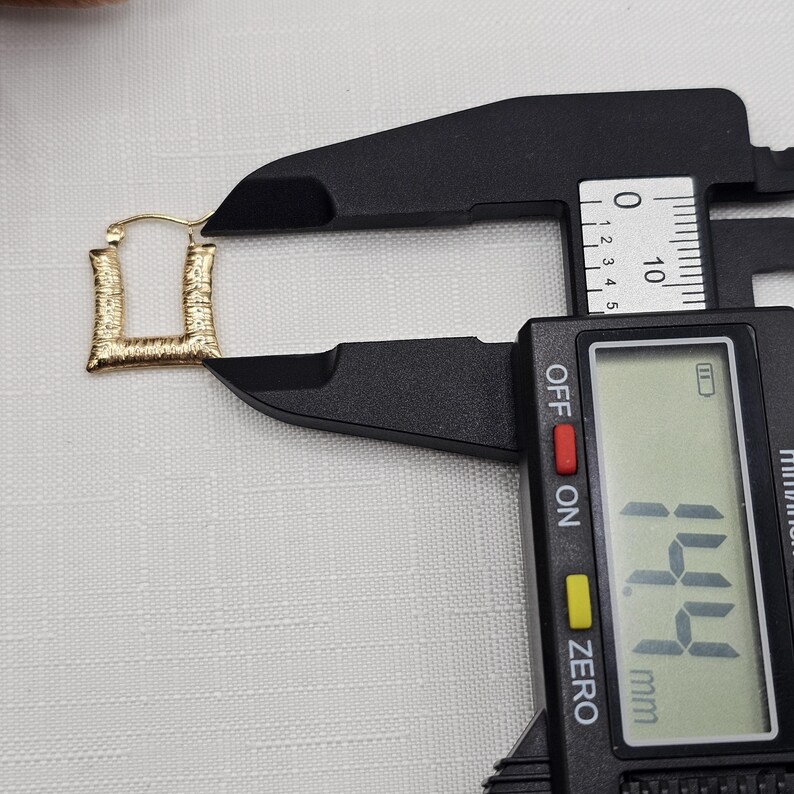 May include: A gold-colored, square-shaped earring is measured with a black digital caliper. The earring has a textured surface. The caliper displays a measurement of 14.4 mm.