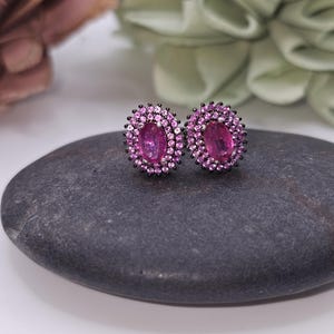 May include: A pair of oval-shaped pink gemstone stud earrings with a halo of smaller pink stones and black metal settings. The earrings are displayed on a dark gray stone, with blurred floral accents in the background.