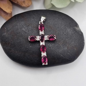 May include: A silver pendant in the shape of a cross, adorned with oval-cut, deep red gemstones and small, clear accent stones. The cross is displayed on a dark, textured surface, showcasing the jewelry's design and color.