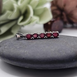 May include: A silver pendant featuring five oval-cut, deep red gemstones. The gemstones are set in a row, creating a linear design. The pendant is displayed on a gray stone, with a blurred background of greenery and brown accents.
