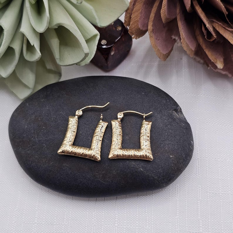 May include: A pair of gold-colored, textured square hoop earrings. The earrings are displayed on a dark gray stone, with a blurred background featuring decorative elements in shades of green and brown.