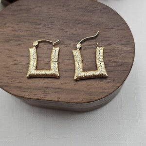 May include: A pair of gold-colored square hoop earrings. The earrings have a textured surface and a post closure. The earrings are displayed on a dark wooden surface.