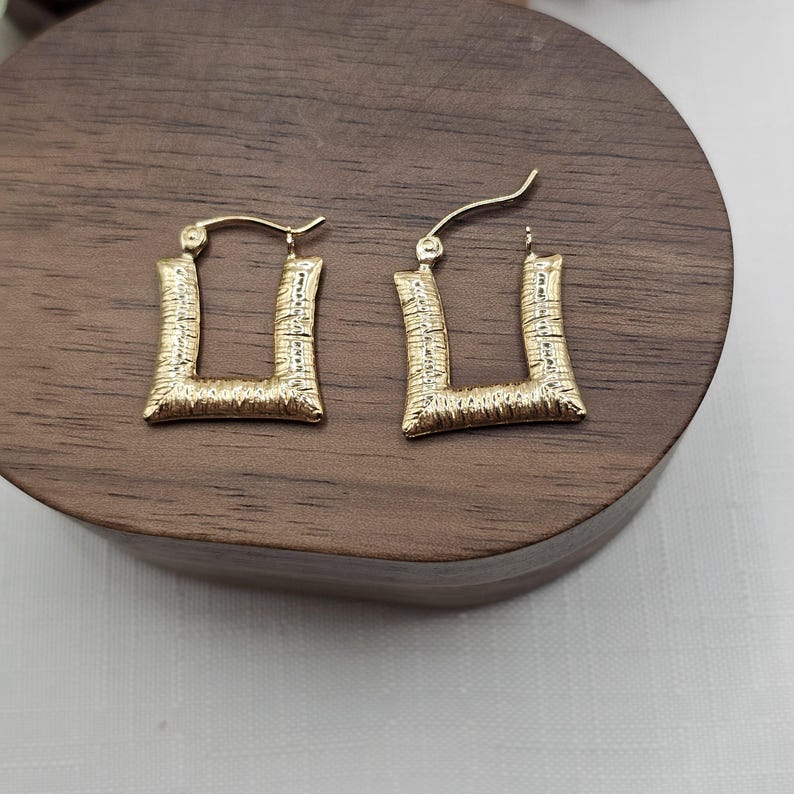May include: A pair of gold-colored square hoop earrings. The earrings have a textured surface and a post closure. The earrings are displayed on a dark wooden surface.