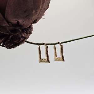 May include: A pair of gold-colored, square-shaped earrings with a textured surface. The earrings are hanging from a green string against a white background. A dried, dark brown flower is visible in the upper left corner.