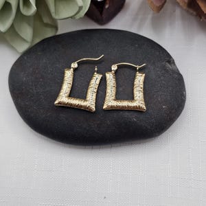 May include: A pair of gold-colored, square hoop earrings. The earrings have a textured surface and a hinged closure. The earrings are displayed on a black stone surface.