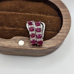 May include: A pair of silver earrings with a vertical design. Each earring features a row of oval-cut, deep red gemstones, alternating with smaller, clear gemstones. The earrings are set against a wooden jewelry box with a brown interior.