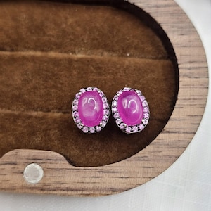 May include: A pair of oval-shaped stud earrings. Each earring features a large, vibrant pink gemstone surrounded by a halo of smaller pink stones. The earrings are set in a silver-colored metal and rest on a brown, velvet-lined surface.