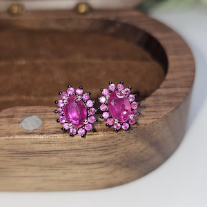 May include: A pair of oval-shaped, magenta gemstone stud earrings. Each earring features a large central stone surrounded by smaller, round magenta stones. The settings are black, and the earrings are displayed on a wooden surface.