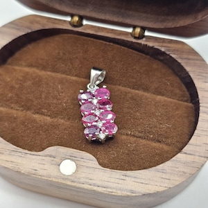 May include: A silver pendant featuring a vertical arrangement of oval-cut, vibrant pink gemstones, interspersed with smaller, clear stones. The pendant is displayed inside a wooden jewelry box with a brown velvet lining.