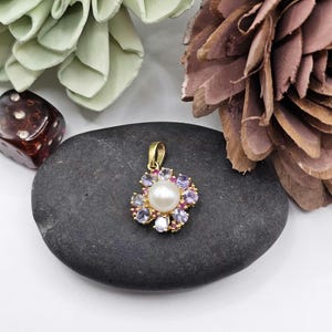 May include: A gold pendant with a flower design, featuring a central white pearl surrounded by purple and red gemstones. The pendant is set on a dark gray stone, with decorative elements in the background.