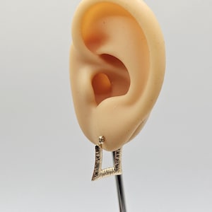 May include: A gold-colored square earring with a textured surface is shown. The earring is attached to a post. The earring is displayed next to a mannequin ear. The background is white.
