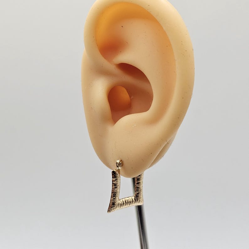 May include: A gold-colored square earring with a textured surface is shown. The earring is attached to a post. The earring is displayed next to a mannequin ear. The background is white.