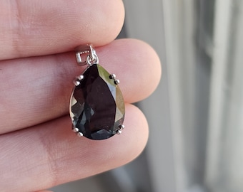 May include: A teardrop-shaped pendant featuring a faceted dark black stone set in a silver-toned metal prong setting. The pendant includes a small silver-toned metal bail at the top for attachment to a chain or cord, showcasing a polished finish.