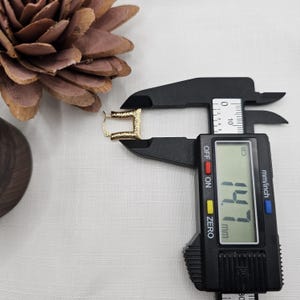 May include: A gold-colored, textured rectangular earring is measured with a black digital caliper. The caliper displays a measurement of 14.7 mm. A brown pinecone and a wooden object are in the background.