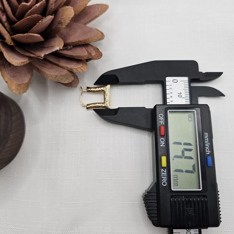 May include: A gold-colored, textured rectangular earring is measured with a black digital caliper. The caliper displays a measurement of 14.7 mm. A brown pinecone and a wooden object are in the background.