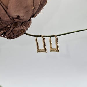 May include: A pair of gold-colored, rectangular earrings with a textured surface. The earrings are hanging from a thin, green stem against a soft, white background. The earrings have a simple, geometric design.