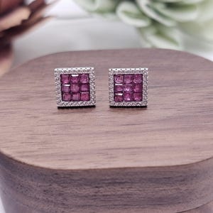 May include: A pair of square stud earrings. Each earring features a grid of pink gemstones surrounded by a border of small, clear stones. The earrings are set on a silver-colored metal. The background is a wooden surface.