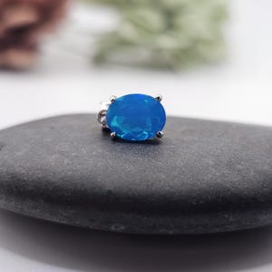 May include: A vibrant blue gemstone pendant with a silver setting. The oval-shaped stone is a bright, eye-catching blue. The pendant is displayed on a dark gray stone, with a blurred background of green and brown.