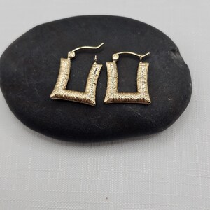 May include: A pair of gold-colored square hoop earrings with a textured surface. The earrings are displayed on a dark, oval-shaped stone against a white background. The earrings have a post and hook closure.