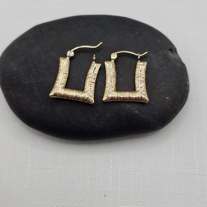 May include: A pair of gold-colored square hoop earrings with a textured surface. The earrings are displayed on a dark, oval-shaped stone against a white background. The earrings have a post and hook closure.