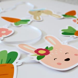 Puede incluir: Una guirnalda de Pascua en colores pastel con recortes de conejos y zanahorias. Los recortes de conejos son rosas con una flor y las zanahorias son naranjas con hojas verdes. La guirnalda está ensartada en una cinta blanca.