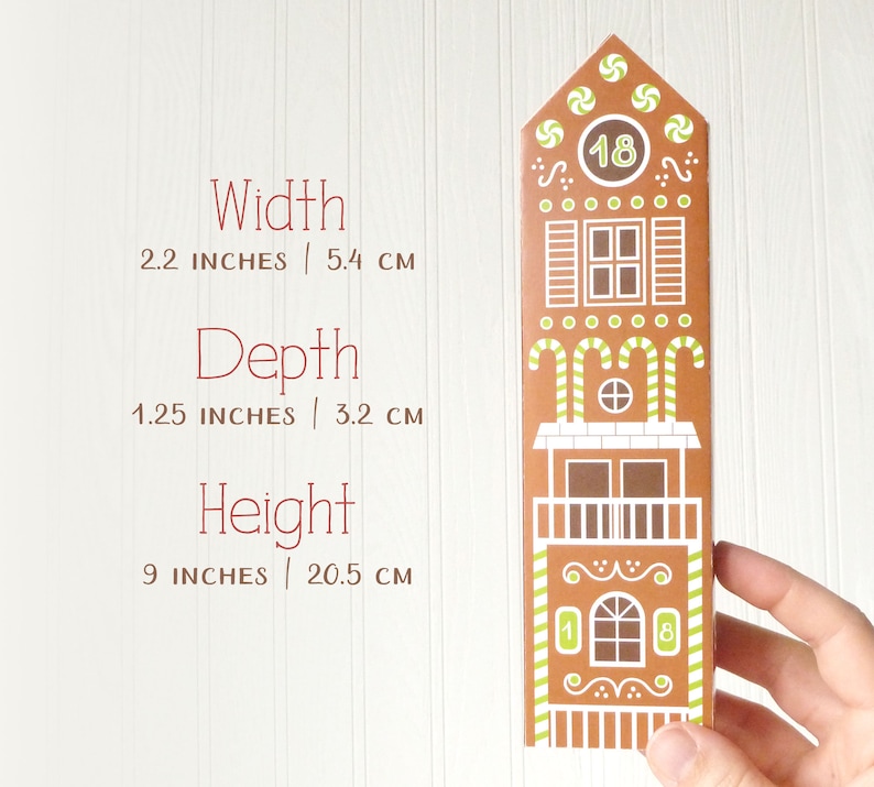 Hand holding a finished advent calendar gingerbread house to demonstrate it's relative size: 2.2 by 1.25 by 9 inches, or 5.4 by 3.2 by 20.5 cm