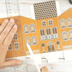 May include: A person is assembling a brown and white paper gingerbread house. The house has a white picket fence, a brown door, and white windows. The person is using a ruler and a craft knife to cut the paper.
