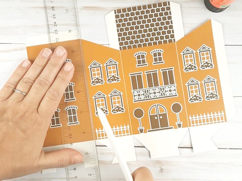 May include: A person is assembling a brown and white paper gingerbread house. The house has a white picket fence, a brown door, and white windows. The person is using a ruler and a craft knife to cut the paper.