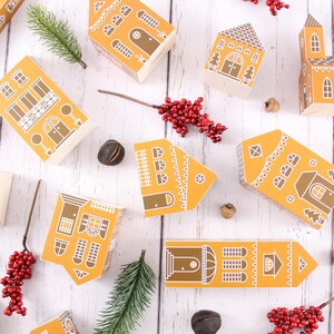 May include: A collection of 10 small, yellow paper houses with white trim and various designs. The houses are arranged on a white wooden surface with red berries, pine sprigs, and acorns scattered around.