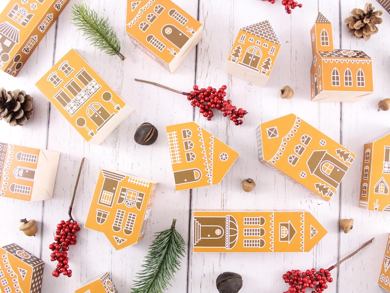 May include: A collection of 10 small, yellow paper houses with white trim and various designs. The houses are arranged on a white wooden surface with red berries, pine sprigs, and acorns scattered around.