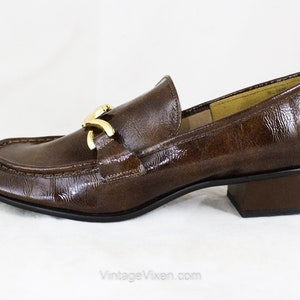 May include: Brown leather heeled loafers with a gold buckle detail. The loafers have a pointed toe and a low block heel.