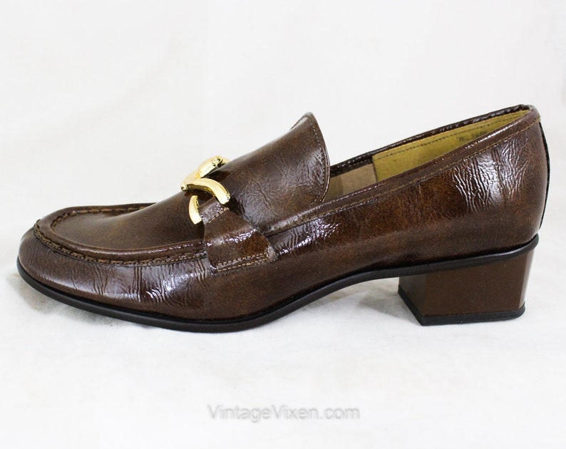 May include: Brown leather heeled loafers with a gold buckle detail. The loafers have a pointed toe and a low block heel.