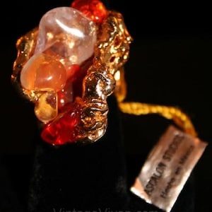 Può includere: Un anello d'oro con un gruppo di pietre rosse, arancioni e trasparenti incastonate in un metallo ruvido e testurizzato. L'anello è etichettato "Asparuh Studio" e "Hand Made".