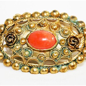Puede incluir: Un broche dorado con una piedra de cabuj&oacute;n de color coral engastada en un dise&ntilde;o de filigrana. El broche est&aacute; rodeado de peque&ntilde;as cuentas redondas y presenta dos acentos en forma de rosa.