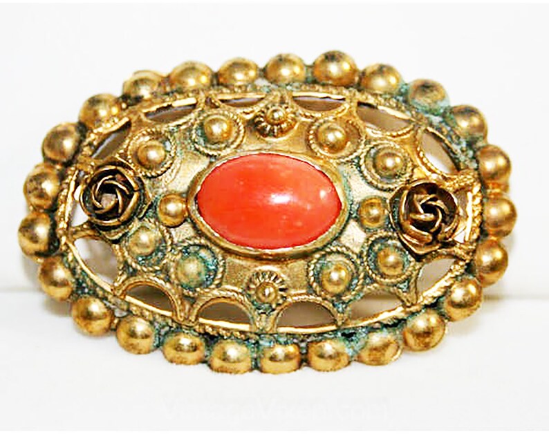 Puede incluir: Un broche dorado con una piedra de cabuj&oacute;n de color coral engastada en un dise&ntilde;o de filigrana. El broche est&aacute; rodeado de peque&ntilde;as cuentas redondas y presenta dos acentos en forma de rosa.