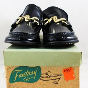 May include: A pair of black leather loafers with gold buckles. The shoes are sitting on a box with the word "Fantasy" printed on it. The box also has a red sale price tag.