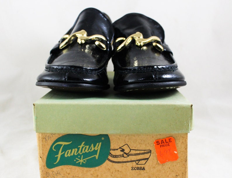 May include: A pair of black leather loafers with gold buckles. The shoes are sitting on a box with the word "Fantasy" printed on it. The box also has a red sale price tag.
