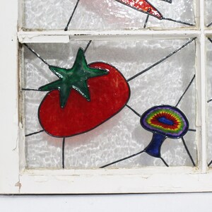 May include: A stained glass window pane featuring a red tomato, a green leaf, and a colourful mushroom. The window pane is set in a white wooden frame.