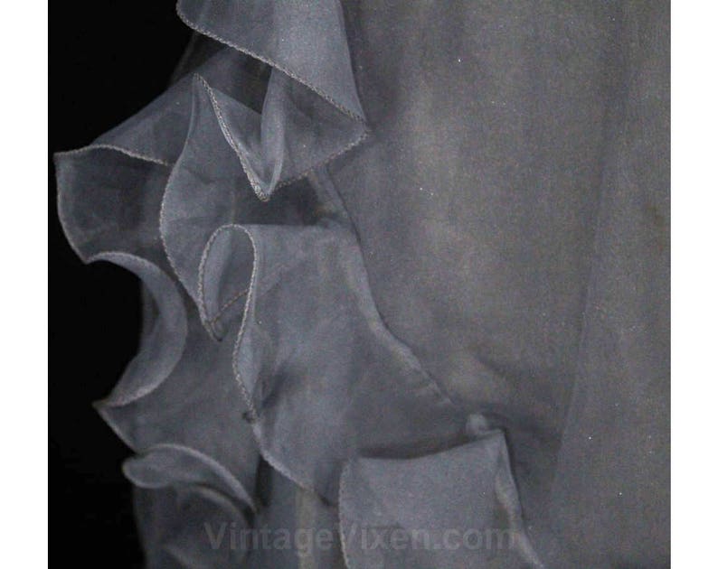 May include: A close-up of a gray tulle fabric with ruffled edges. The fabric is soft and sheer, and the ruffles are delicate and flowing.