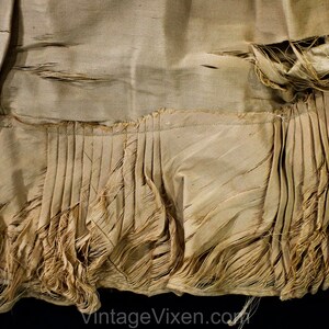 May include: Close-up of a vintage garment in a light beige hue. The fabric features a pleated lower section with frayed edges, showing signs of wear. The texture appears soft and aged, indicating a historical piece of clothing.