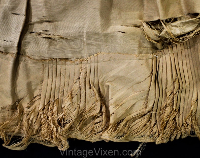 May include: Close-up of a vintage garment in a light beige hue. The fabric features a pleated lower section with frayed edges, showing signs of wear. The texture appears soft and aged, indicating a historical piece of clothing.