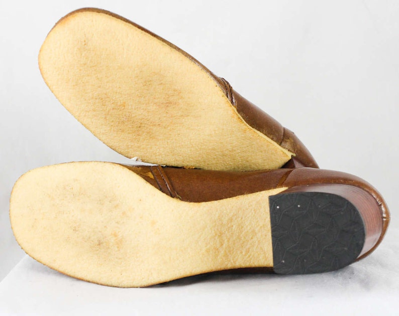 May include: Brown leather heeled boots with a rubber sole. The boots have a brown leather upper and a rubber sole. The boots are shown from the bottom, with the soles facing the camera.