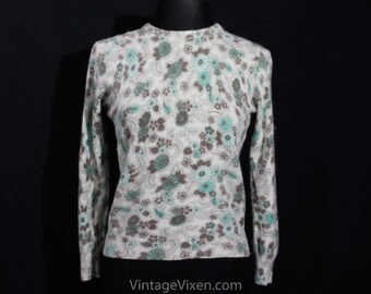 Puede incluir: Un suéter de manga larga con una base de color crema y un patrón floral repetitivo en tonos verde azulado y marrón. La prenda tiene un cuello redondo y una textura suave y difusa en toda su superficie. El texto VintageVixen.com aparece abajo.
